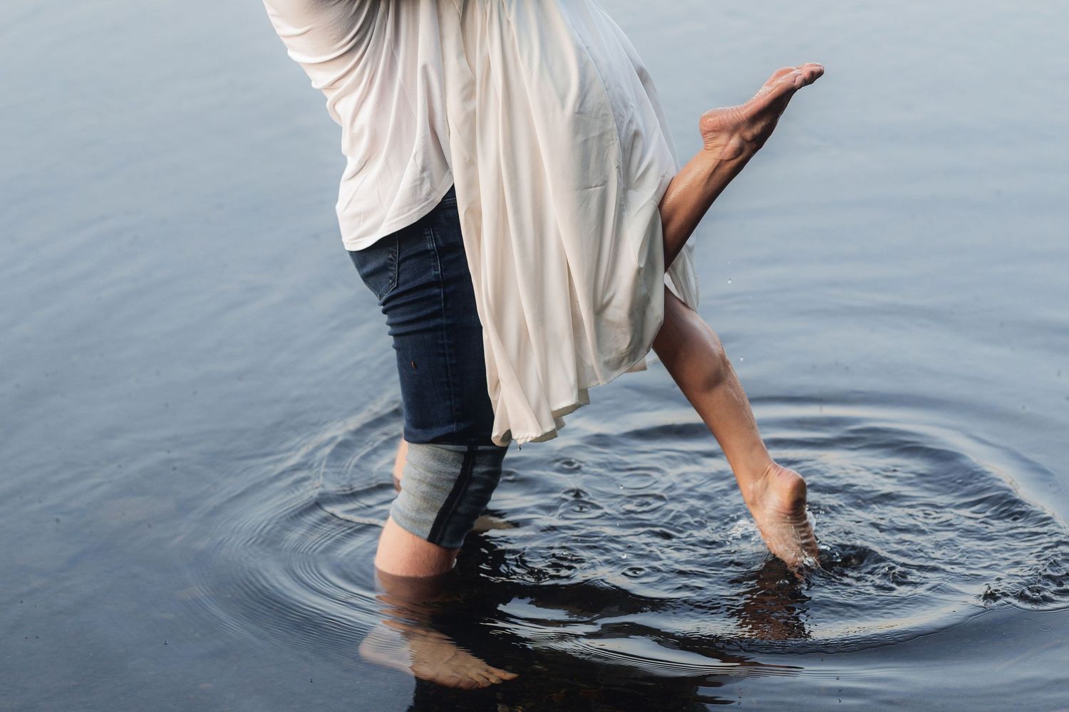 Two people wading barefoot in calm water, wearing jeans and a flowing white top, creating gentle ripples.