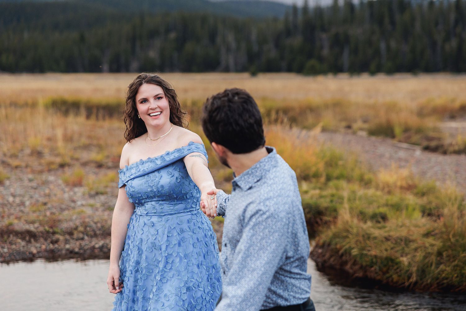 A couple in blue attire poses together against scenic mountain and forest backdrops in a romantic photoshoot series.