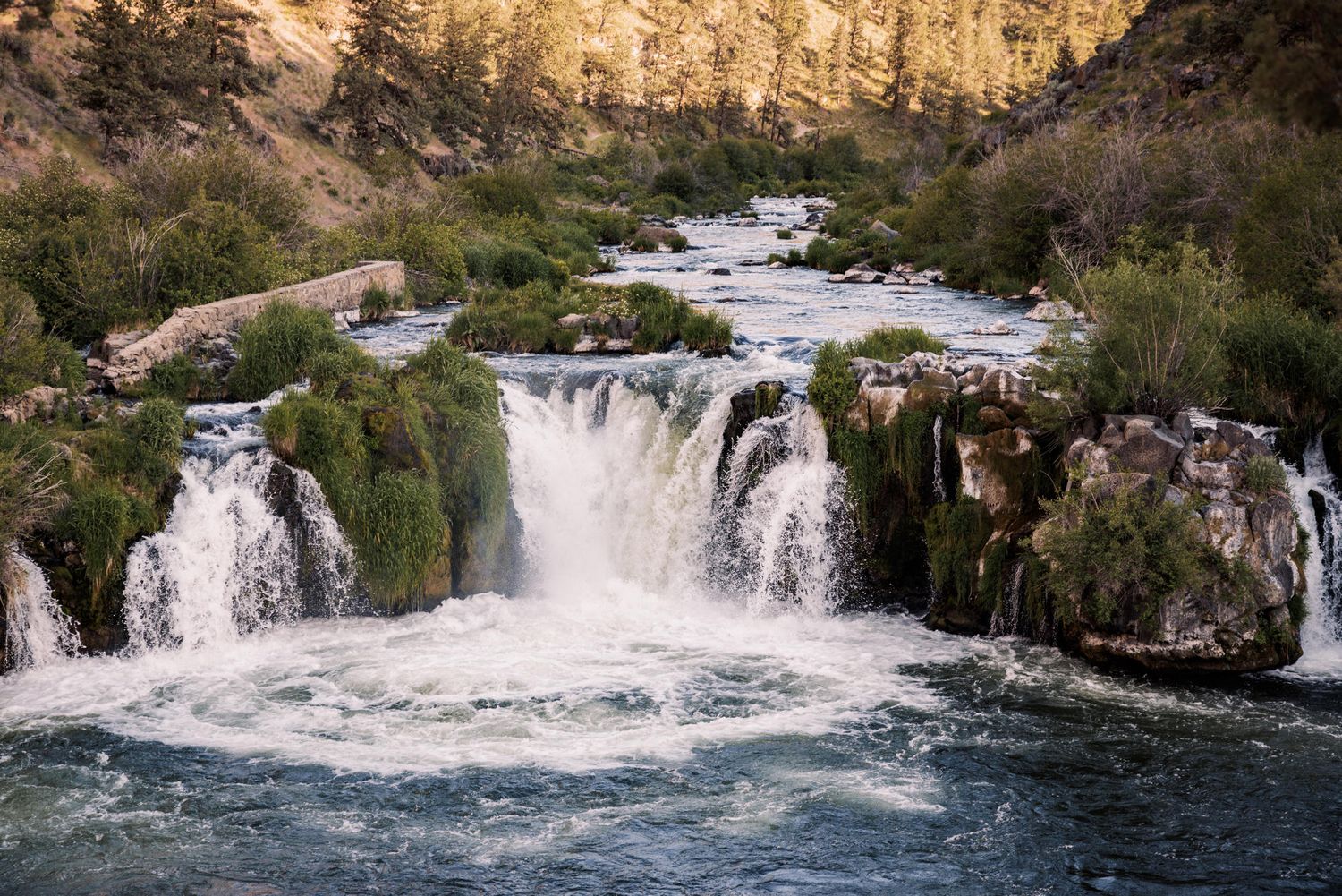 Cascading waterfalls flow over rocky cliffs creating white rapids in a scenic natural landscape during sunset.