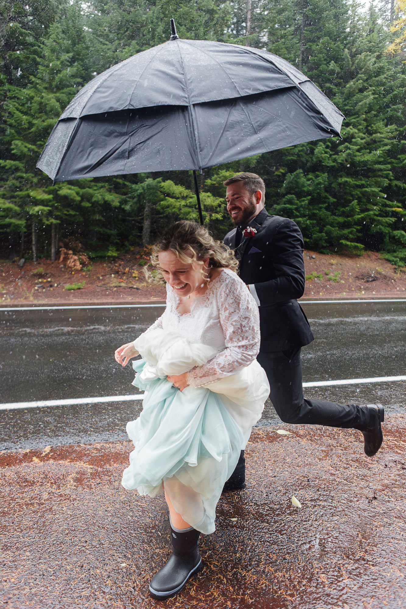 Playful wedding couple splashes through puddles with umbrellas on a rainy day.