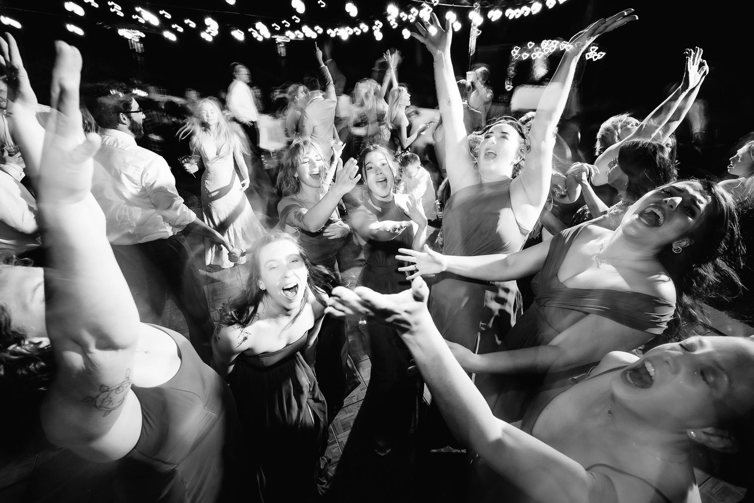 A black and white photo shows a crowded dance floor with people dancing energetically with their hands raised.