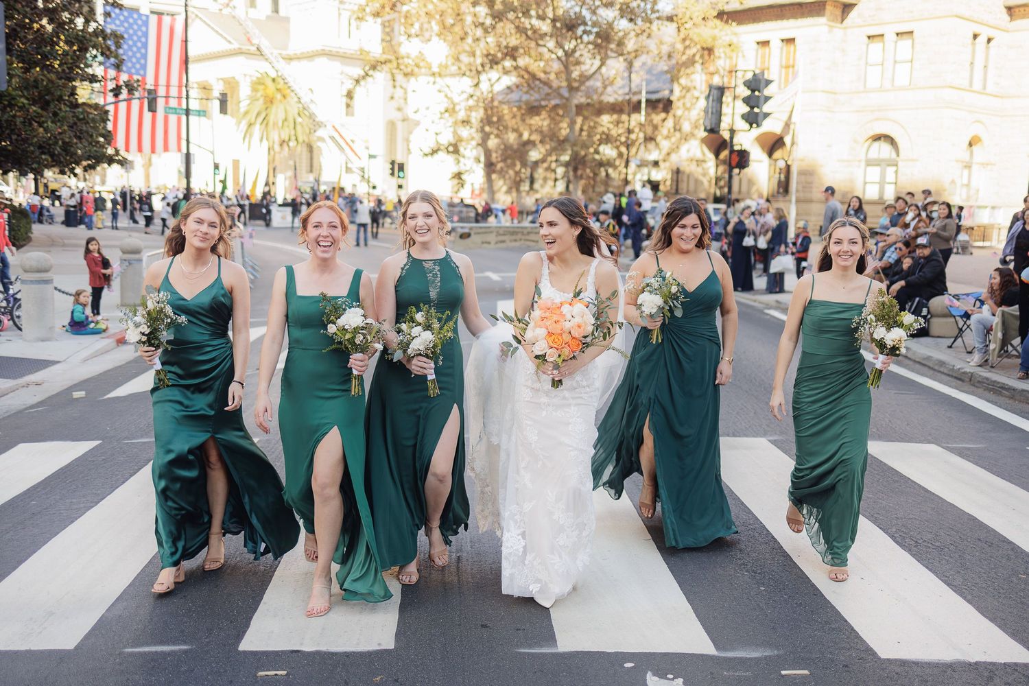 A wedding party in emerald green dresses walks across a crosswalk together during a downtown celebration.