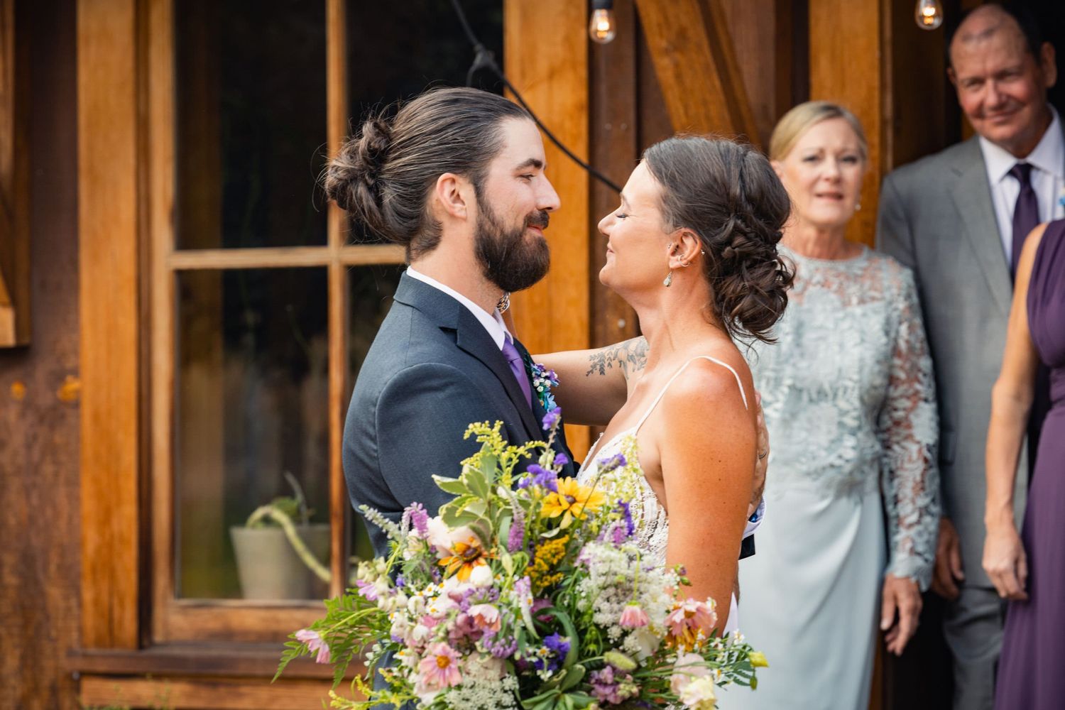 A smiling bride and groom share a joyful moment with wedding guests and bouquet of wildflowers at rustic venue.