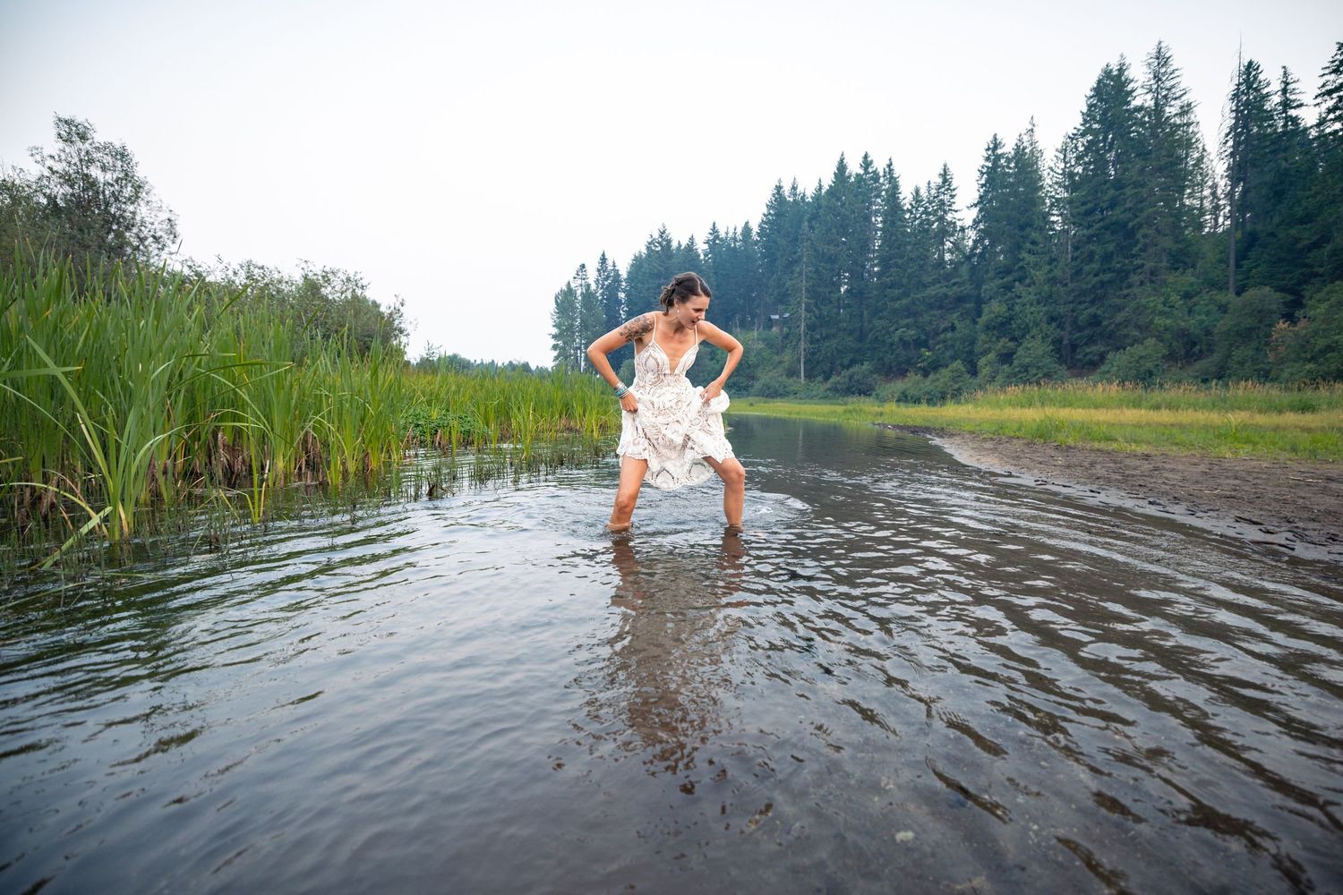 A person in a white dress splashes playfully in shallow lake water surrounded by evergreen trees and reeds.