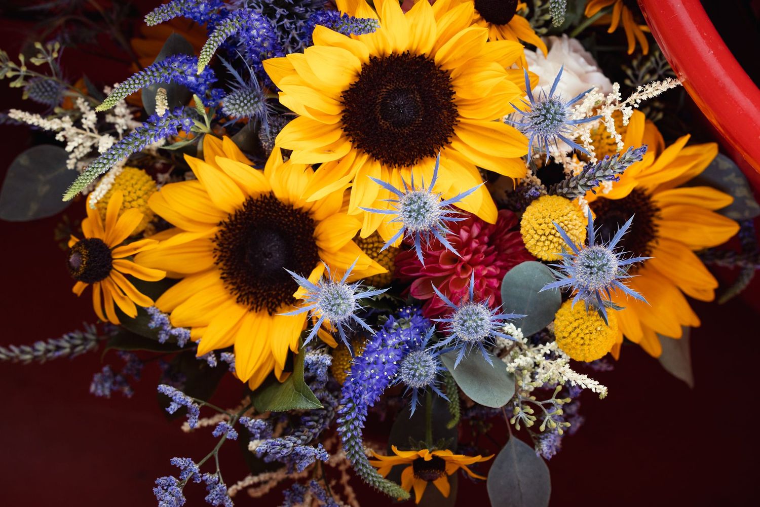 Vibrant floral arrangement featuring bright yellow sunflowers with blue thistles and other wildflowers on a dark surface.