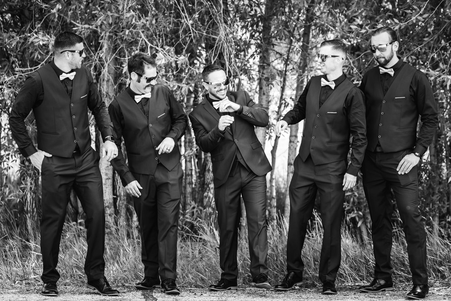 Five men in matching black suits pose together in a dramatic black and white photo against a wooded backdrop.
