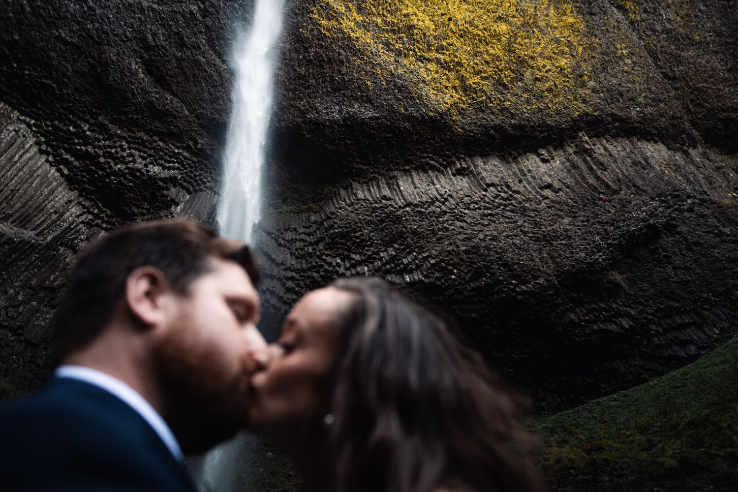 A couple shares a romantic kiss beneath a dramatic waterfall cascading down dark rocky cliffs.