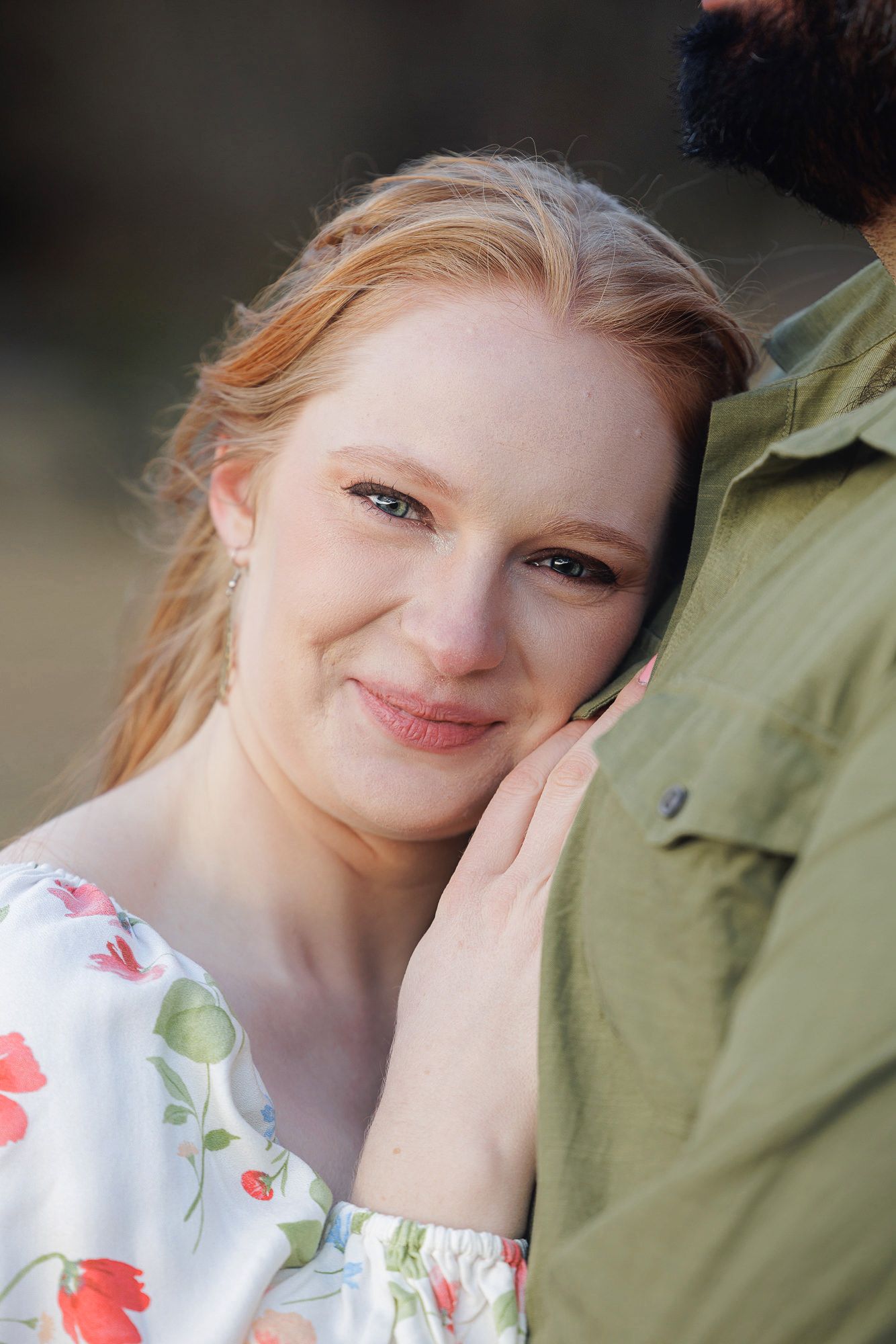 A smiling redhead woman in a floral dress rests her cheek against a man in a green shirt outdoors.