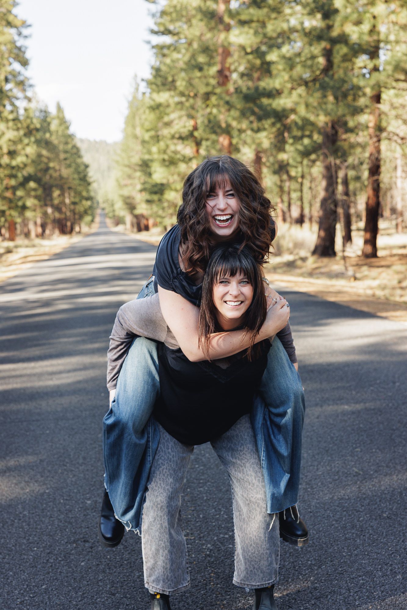 Two laughing women share a joyful piggyback ride on a scenic tree-lined road outdoors.