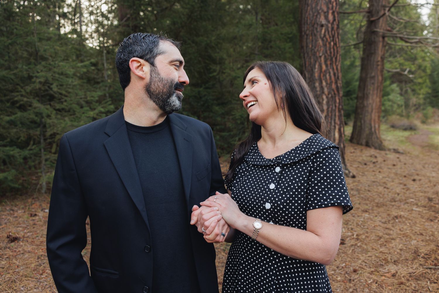 Couple holding hands and smiling at each other among pine trees, man in black suit, woman in polka dot dress.