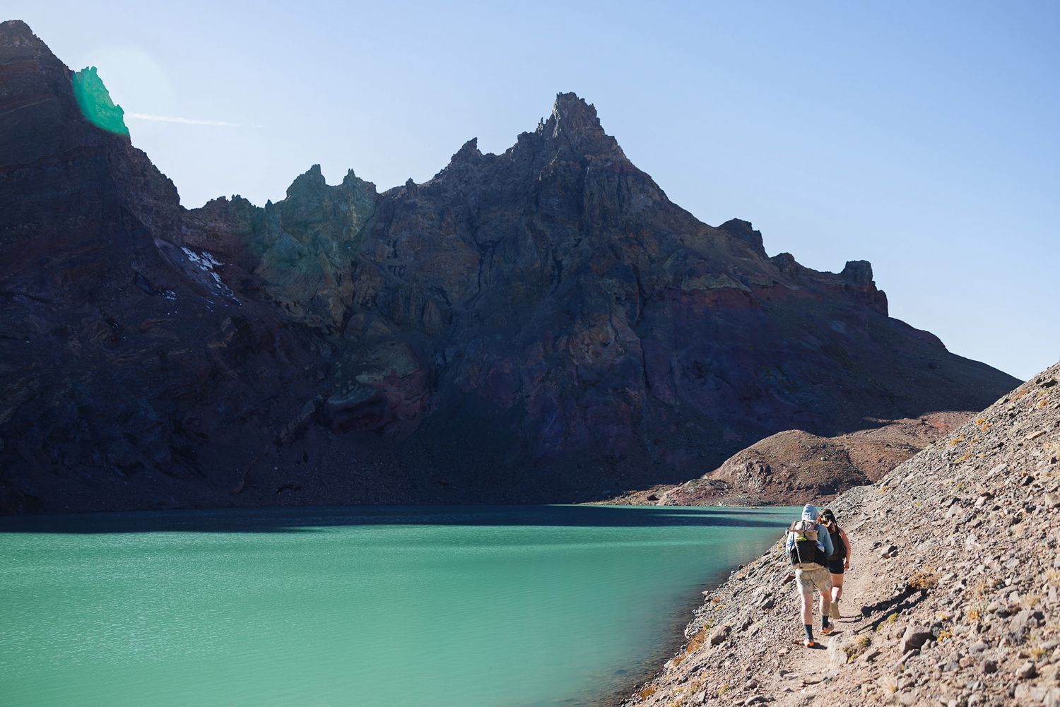 Hiker with a backpack walks along a rocky shoreline beside a turquoise mountain lake beneath rugged dark cliffs and a clear blue sky.