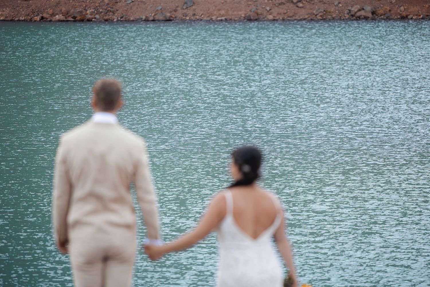 Bride in a white dress and groom in a beige suit hold hands while standing by a sparkling turquoise lake.