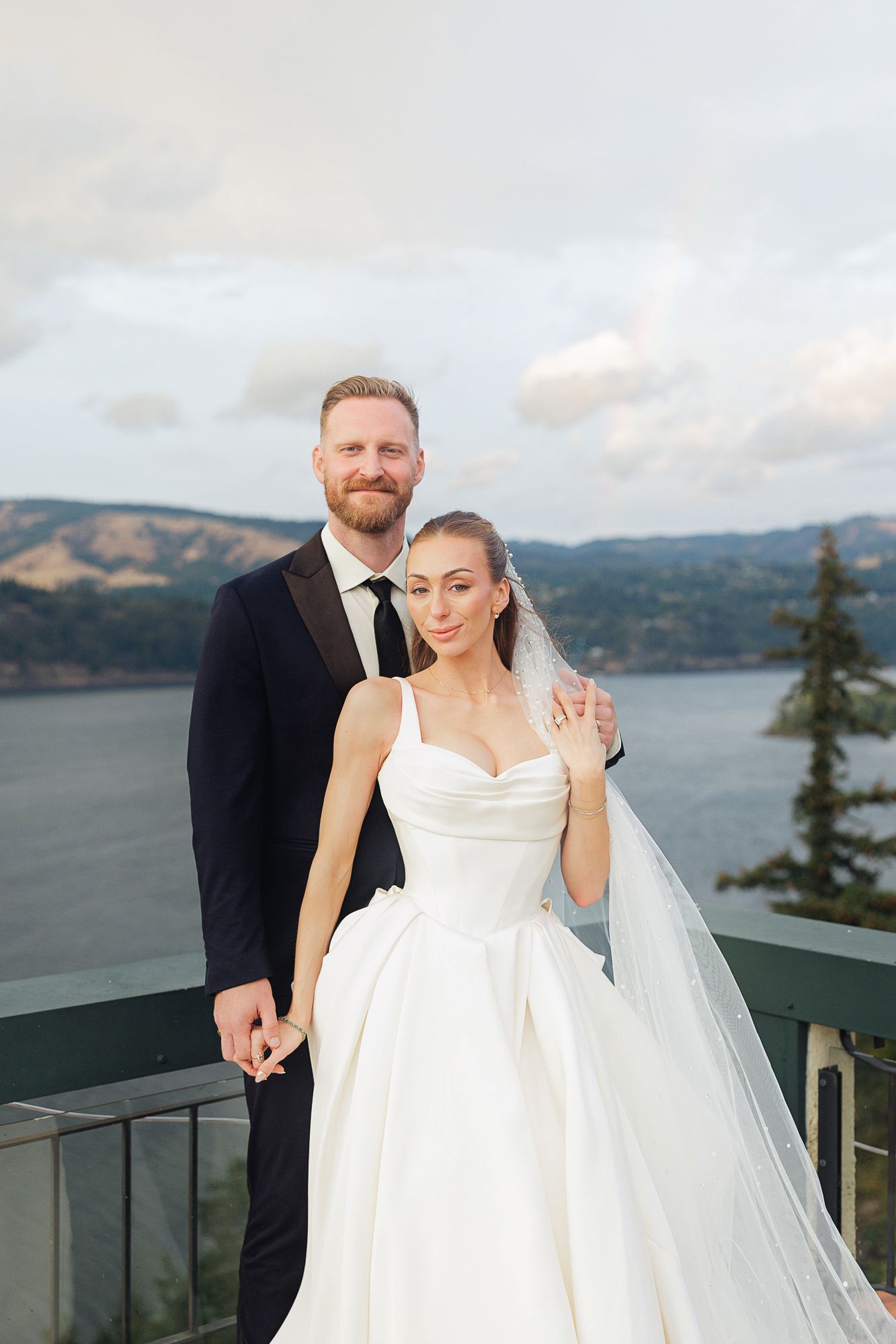Bride in white ball gown and groom in black tuxedo pose on scenic overlook with river and mountains backdrop.