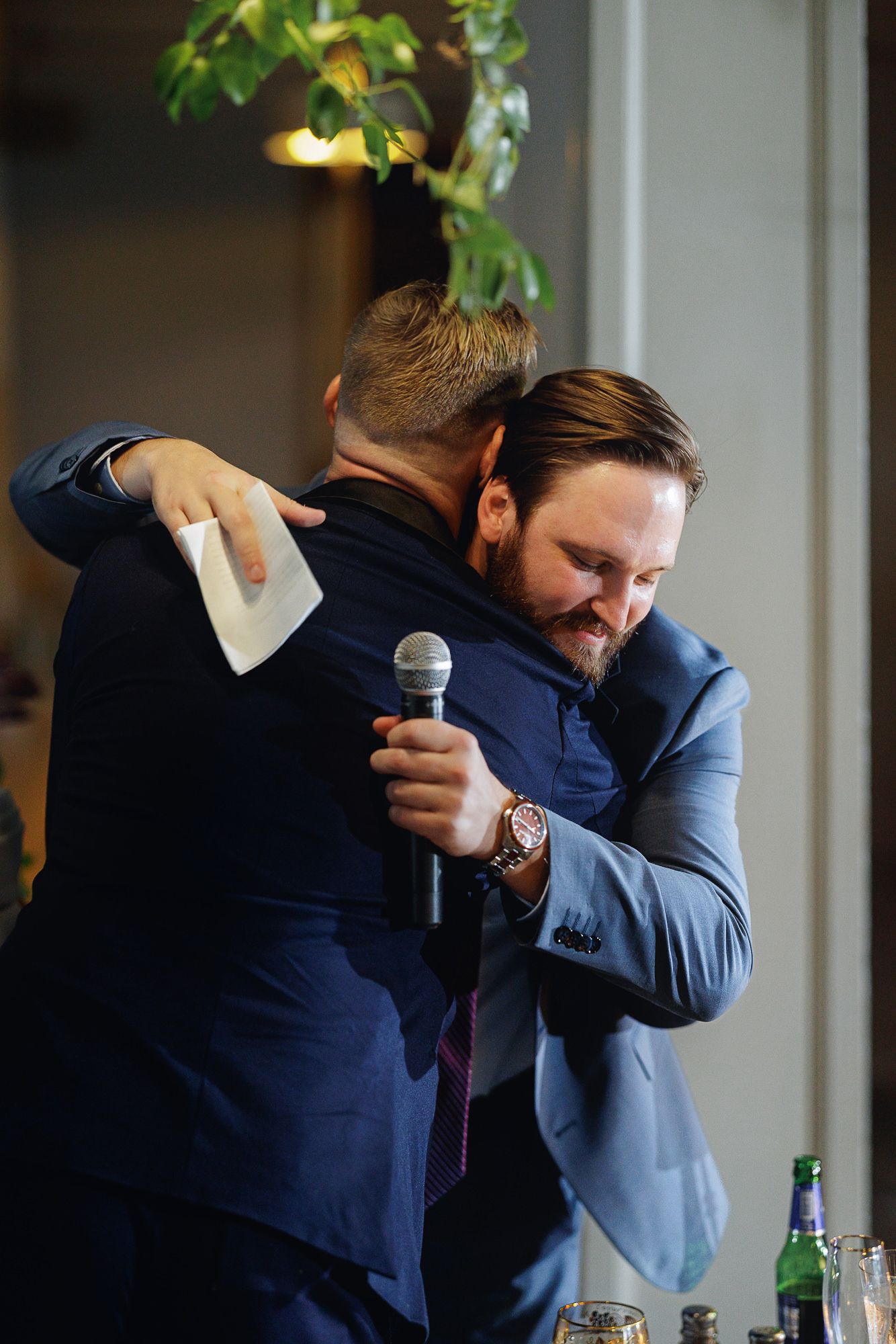 Two men in blue suits share an emotional hug at a wedding reception, one holding a microphone and paper.