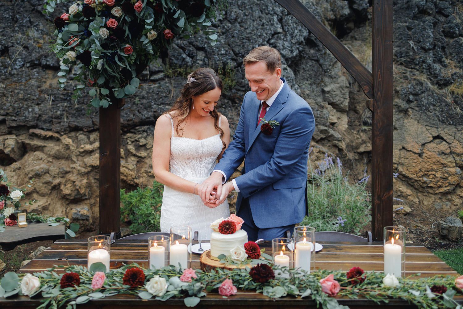 A couple in wedding attire performs a unity candle ceremony at an outdoor venue with rock formations in the background.
