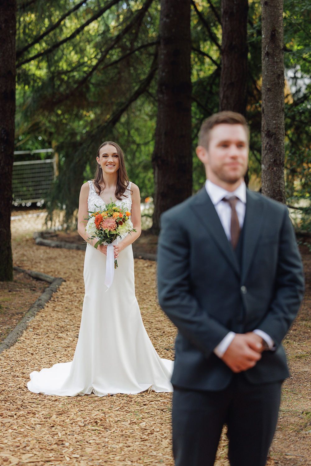 Wedding couple in forest setting with bride holding autumn bouquet and groom in dark suit standing apart.