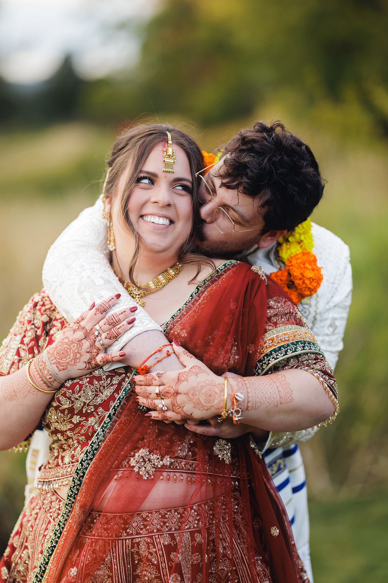 Bride in red lehenga with mehndi hands smiles as groom in sherwani kisses her cheek outdoors.