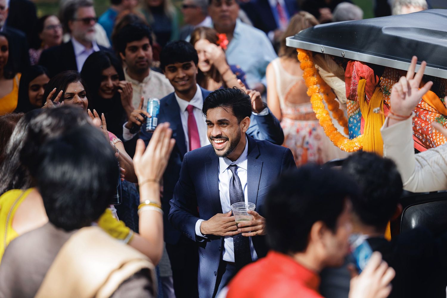 Smiling man in blue suit at vibrant Indian wedding celebration surrounded by guests in colorful attire.