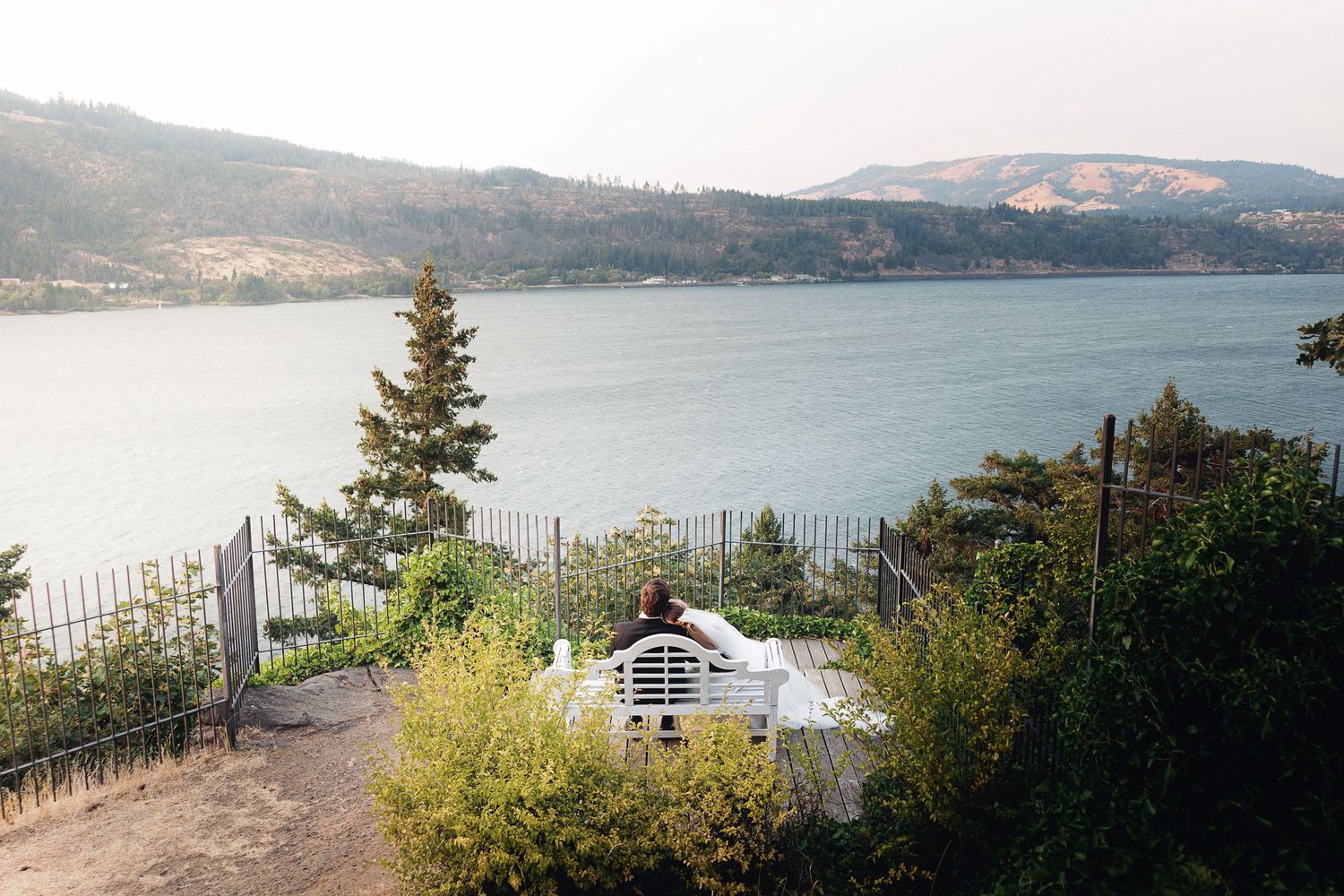 Scenic overlook with white bench facing a serene blue lake surrounded by lush green trees and mountain hills in the background.