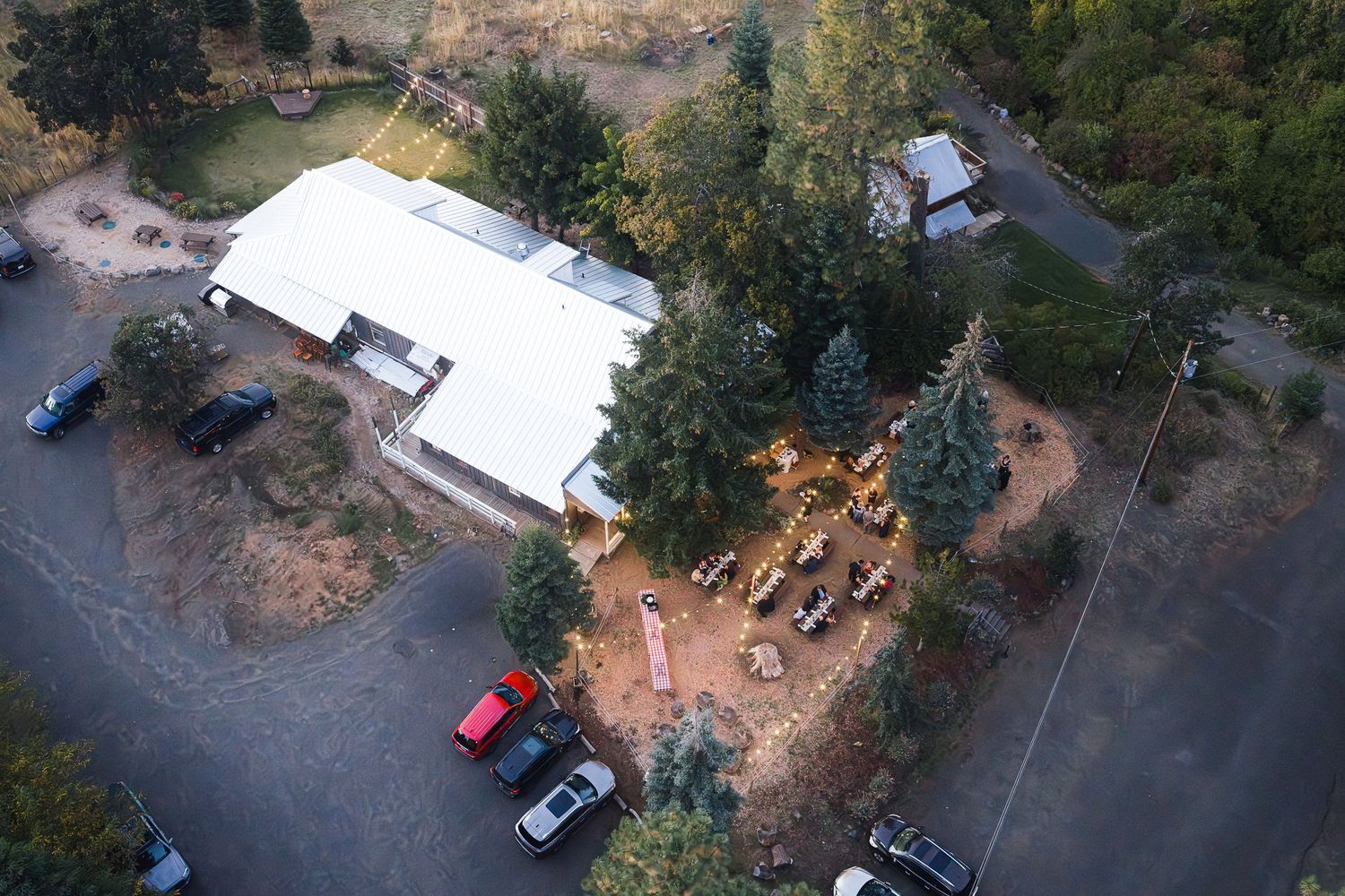 Aerial drone view of a rural property with white building, evergreen trees, and parked cars on a gravel driveway.