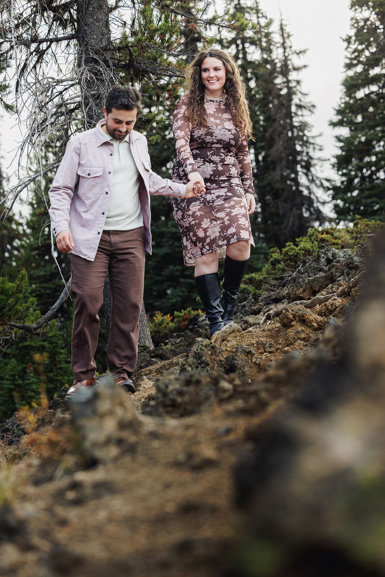 A couple walks together on a rocky mountain trail surrounded by evergreen trees in the Pacific Northwest.