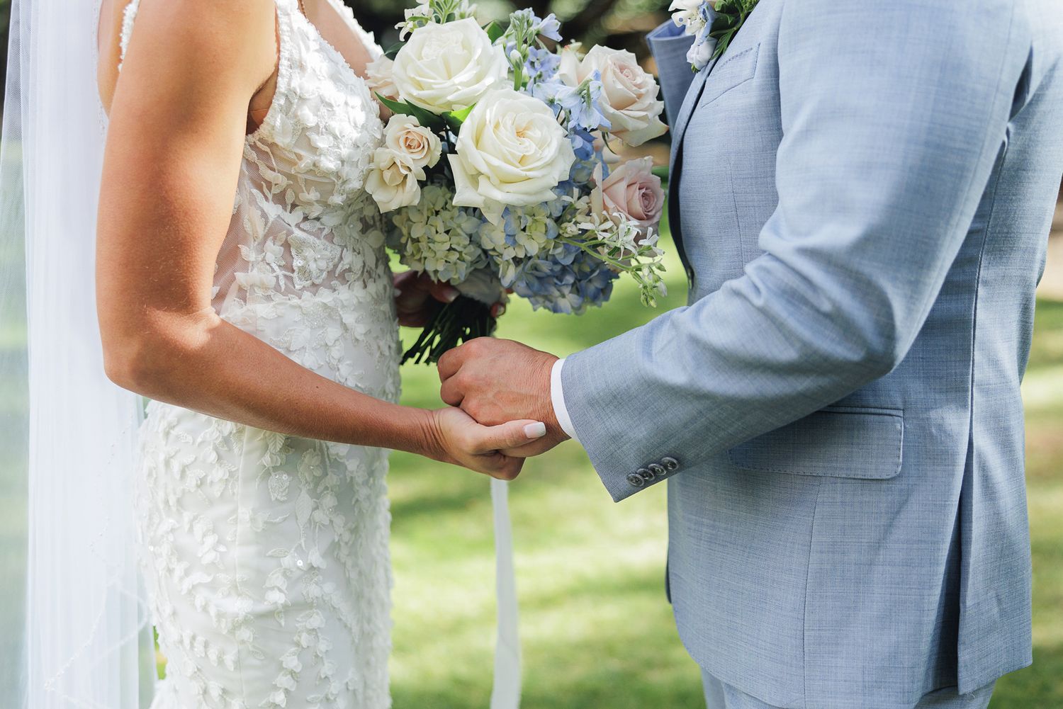 A couple exchanges rings during an outdoor wedding ceremony while holding a bouquet of white roses and greenery.