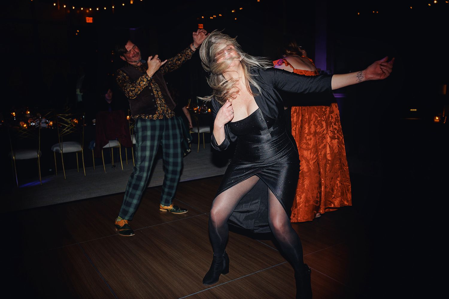 Dancers move energetically on a dark dance floor illuminated by warm string lights overhead.
