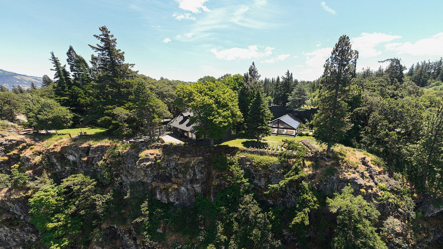 Aerial view of a secluded estate nestled among tall evergreen trees atop rocky cliffs with mountain views.