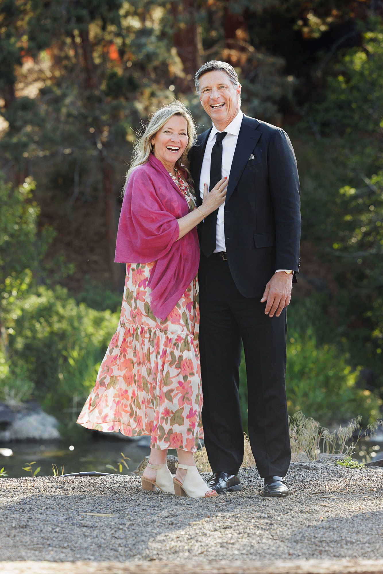 Smiling couple posing outdoors, woman in floral dress with pink shawl, man in black suit, green trees background.