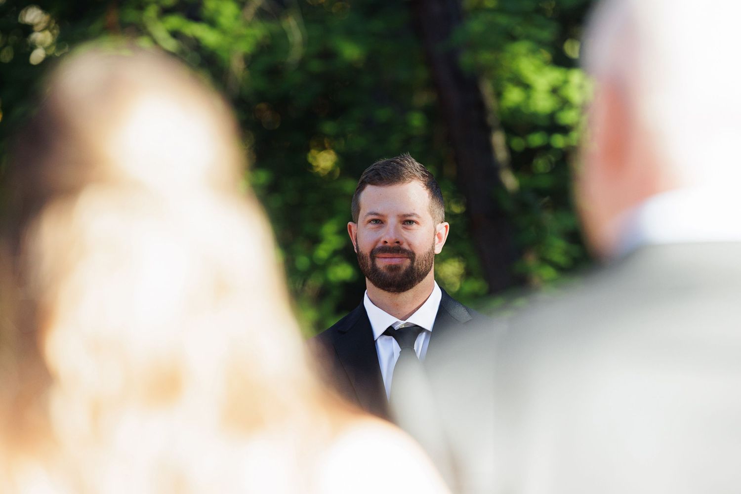 Groom smiling emotionally as bride walks down the aisle during an outdoor wedding ceremony surrounded by lush green trees.