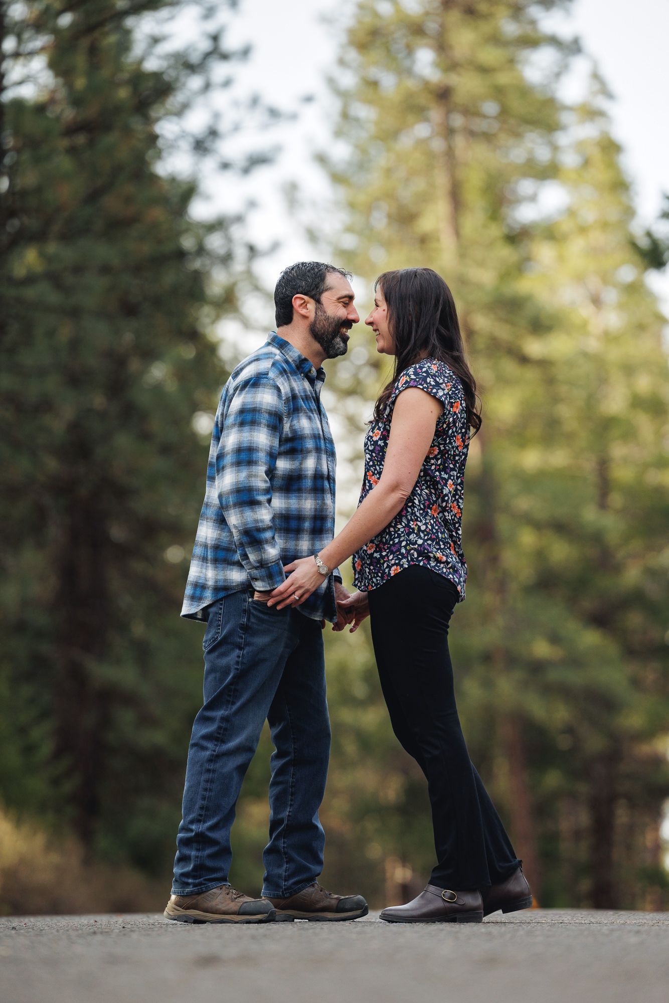 Couple holding hands face to face on a forest path surrounded by tall pine trees during an outdoor engagement session.