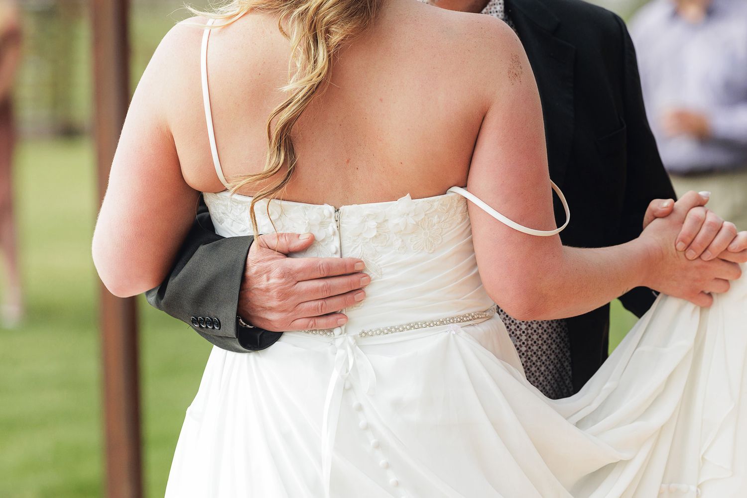 Bride and groom sharing first dance, bride in strapless white wedding gown, groom in dark suit, outdoor ceremony.