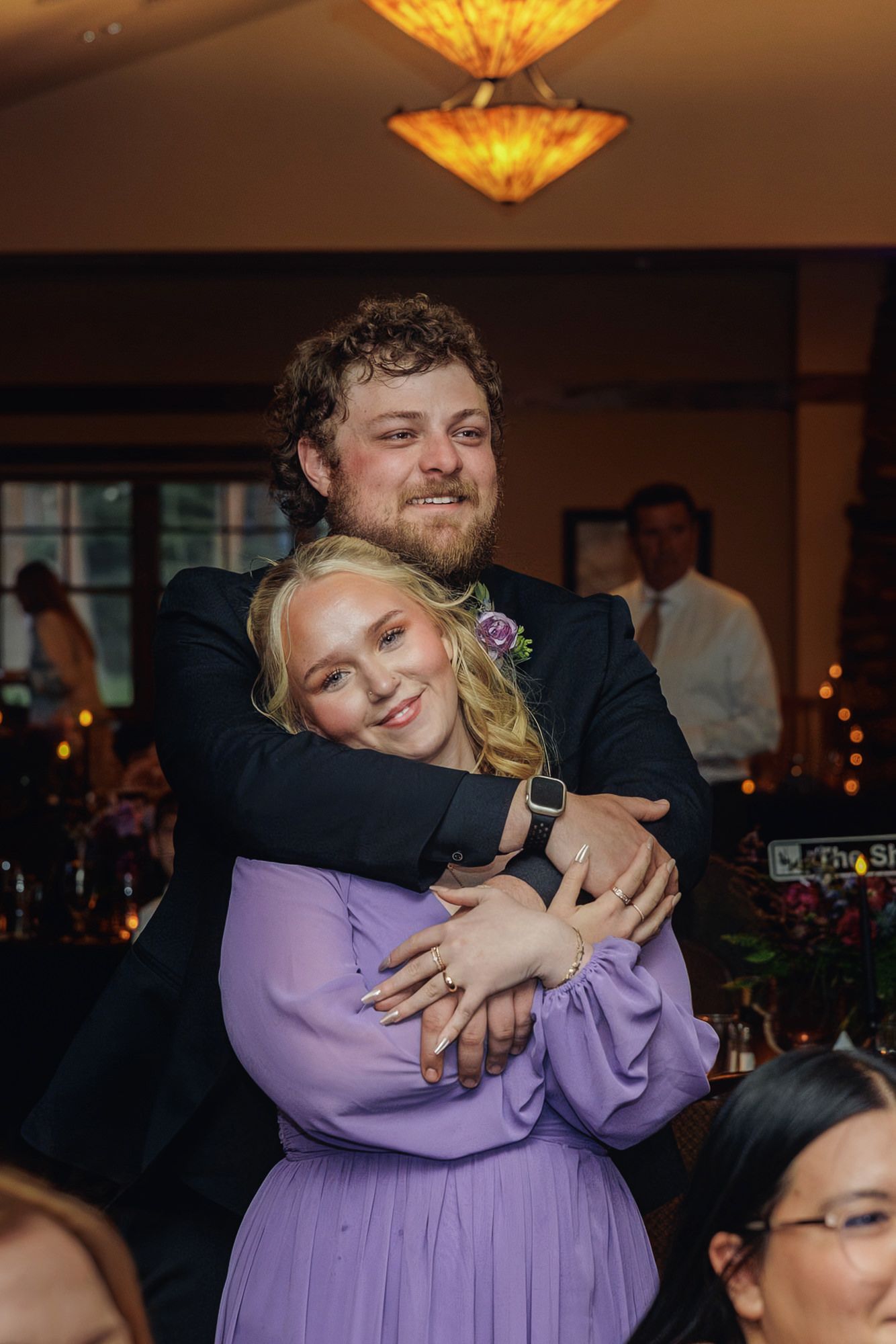 A young woman in a lavender dress hugs a man in a suit at a wedding reception venue.