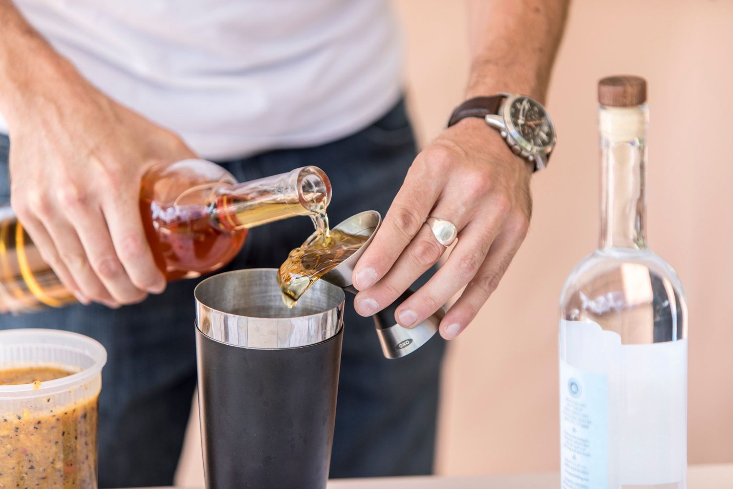 Amber colored liquid being poured into metal cocktail shaker at a bar counter.