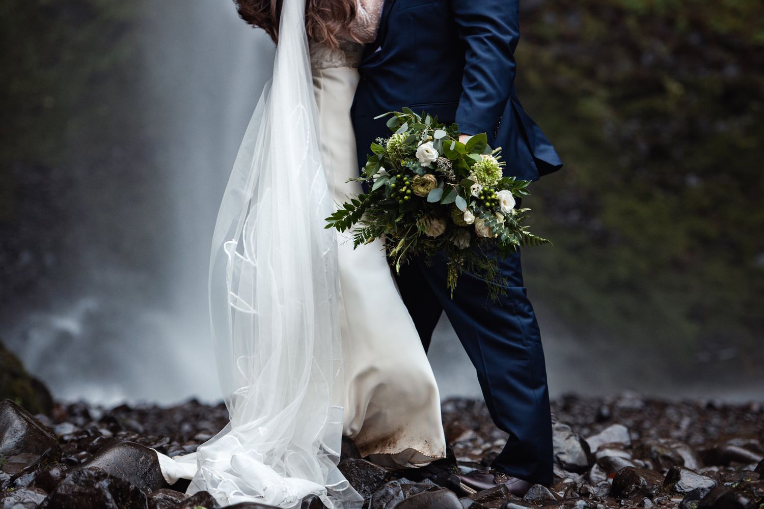 Newlyweds pose near misty waterfall with white dress and blue suit, holding green and white floral bouquet.