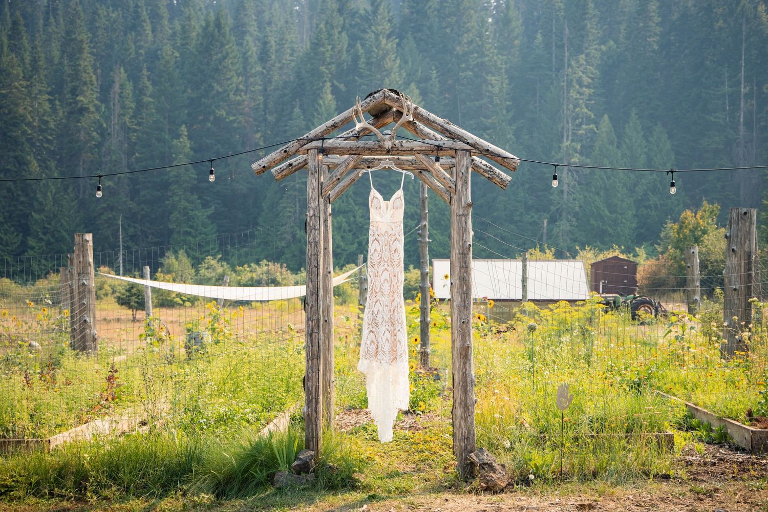 A white wedding dress hangs from a rustic wooden arch in a mountain meadow with evergreen forest in the background.