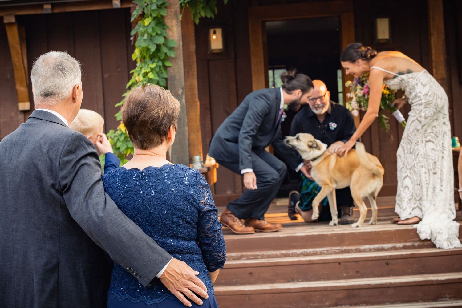 Wedding party gathers on steps to greet an excited dog wearing a bow tie during the celebration.