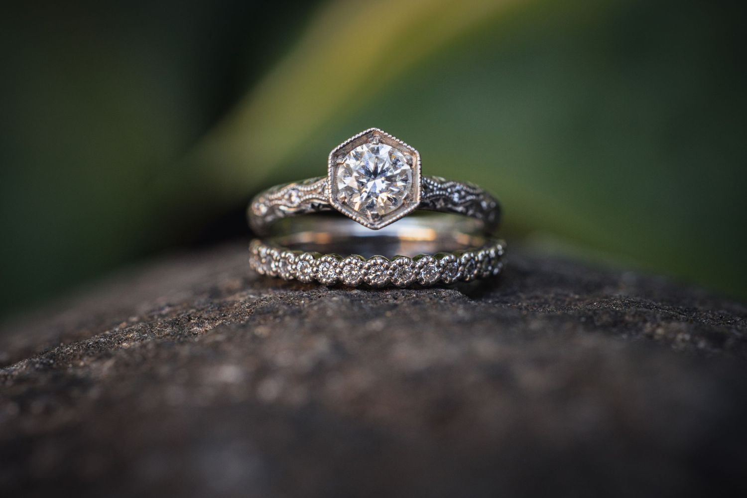 Diamond engagement ring and wedding band displayed on dark stone with blurred green background.
