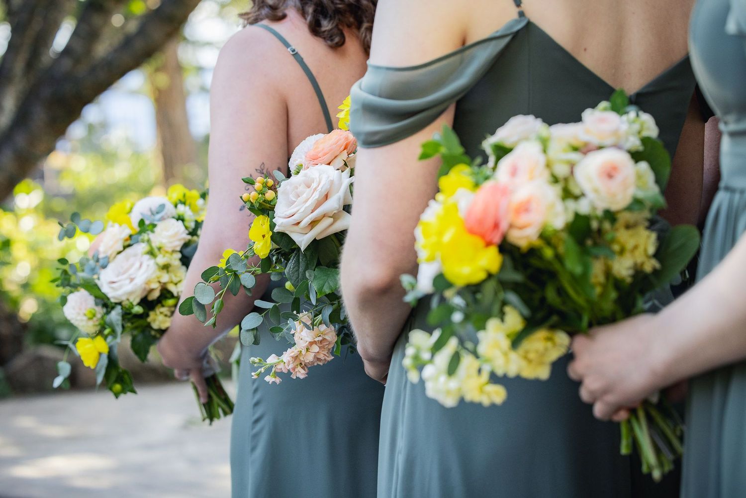 Bridesmaids in sage green dresses hold beautiful bouquets of pink, yellow and white flowers during an outdoor ceremony.