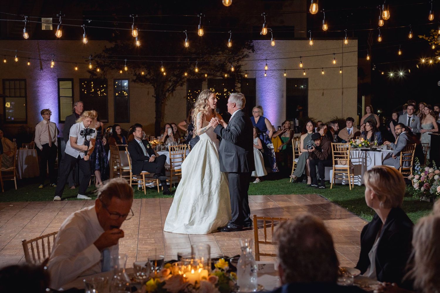 A romantic first dance at an evening wedding reception under glowing string lights and purple uplighting.