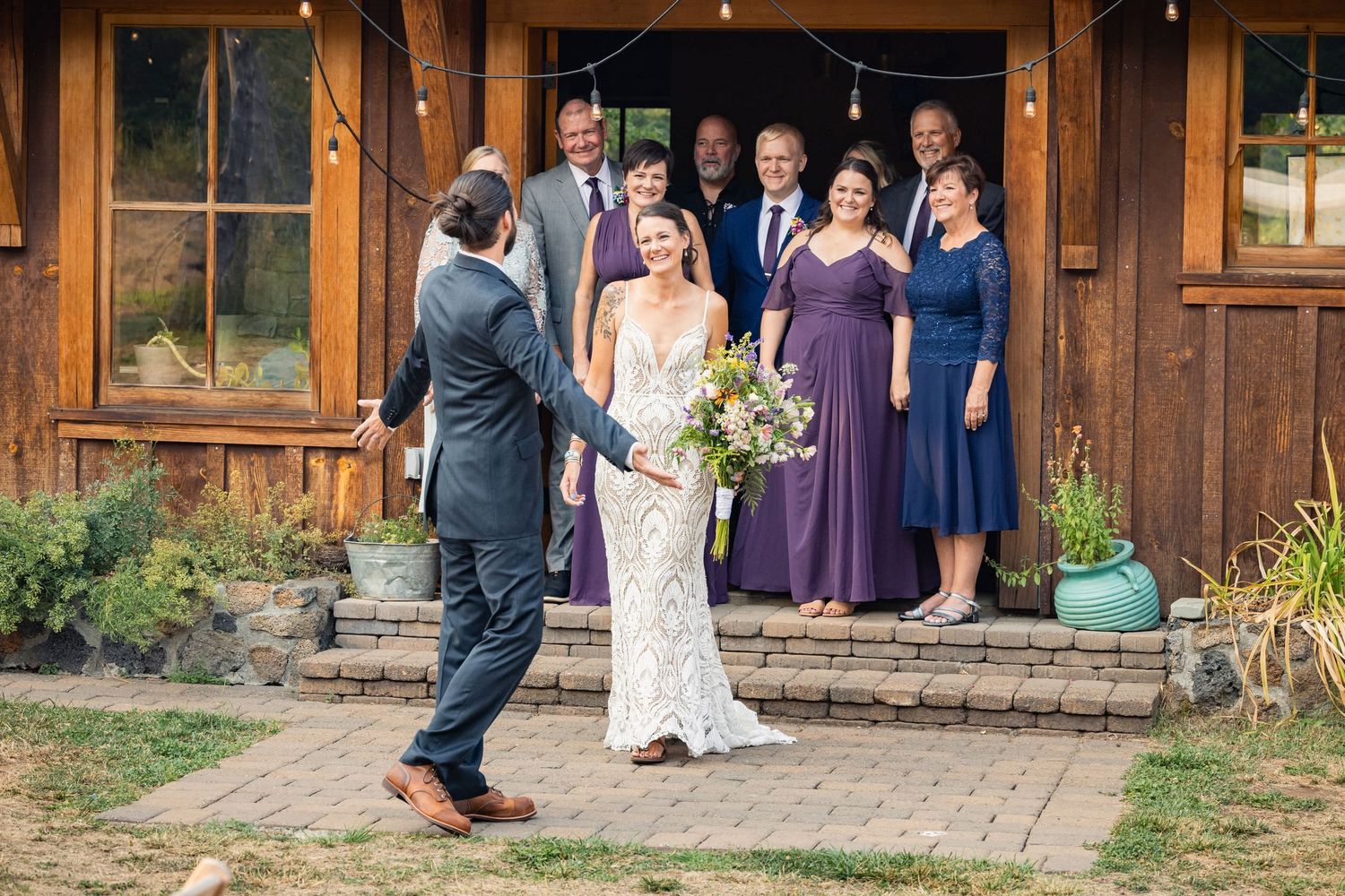 Wedding party stands on rustic cabin porch steps while couple shares a moment on stone pathway.