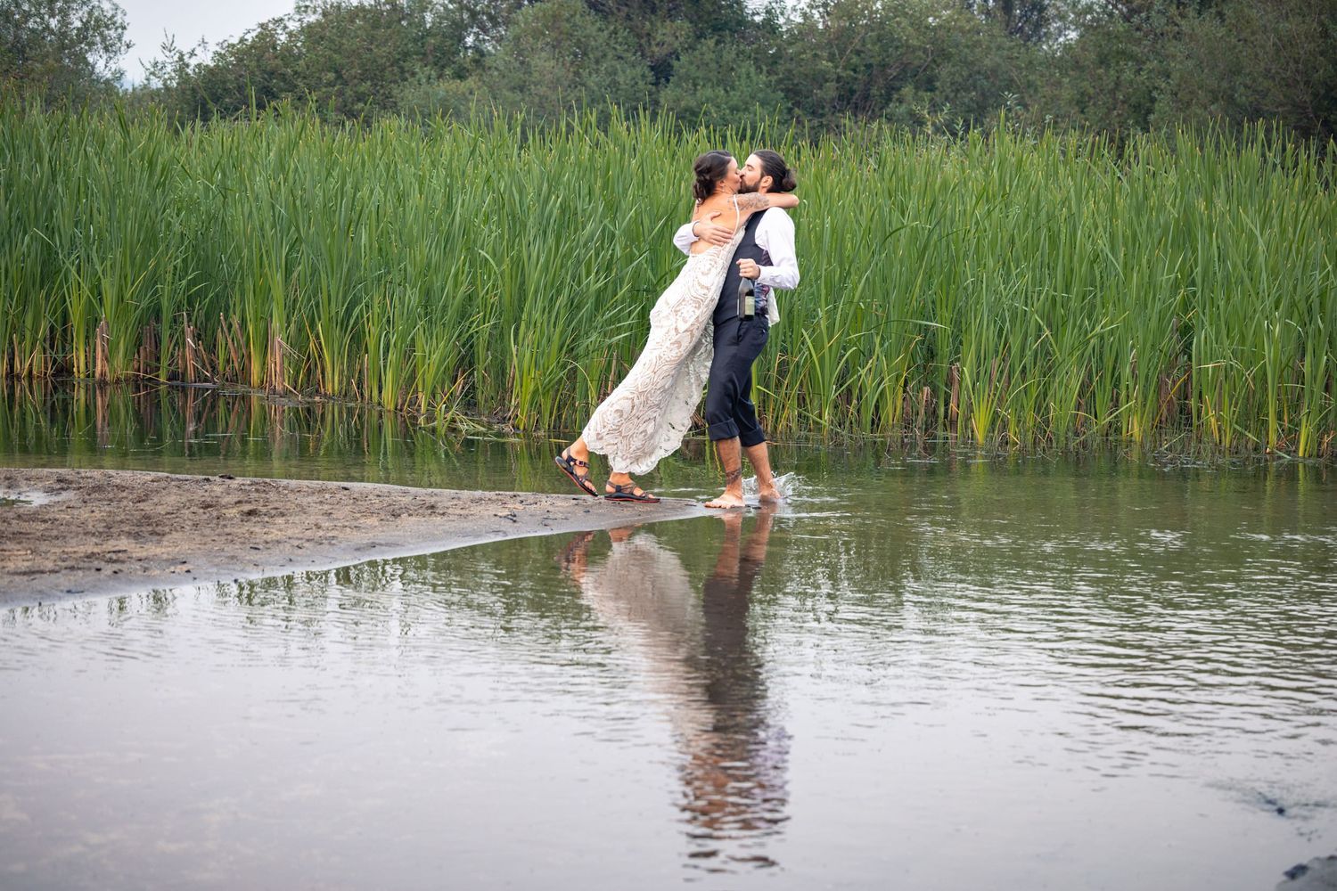 A couple shares a romantic moment on a wooden dock near tall reeds, their reflection showing in the calm water below.