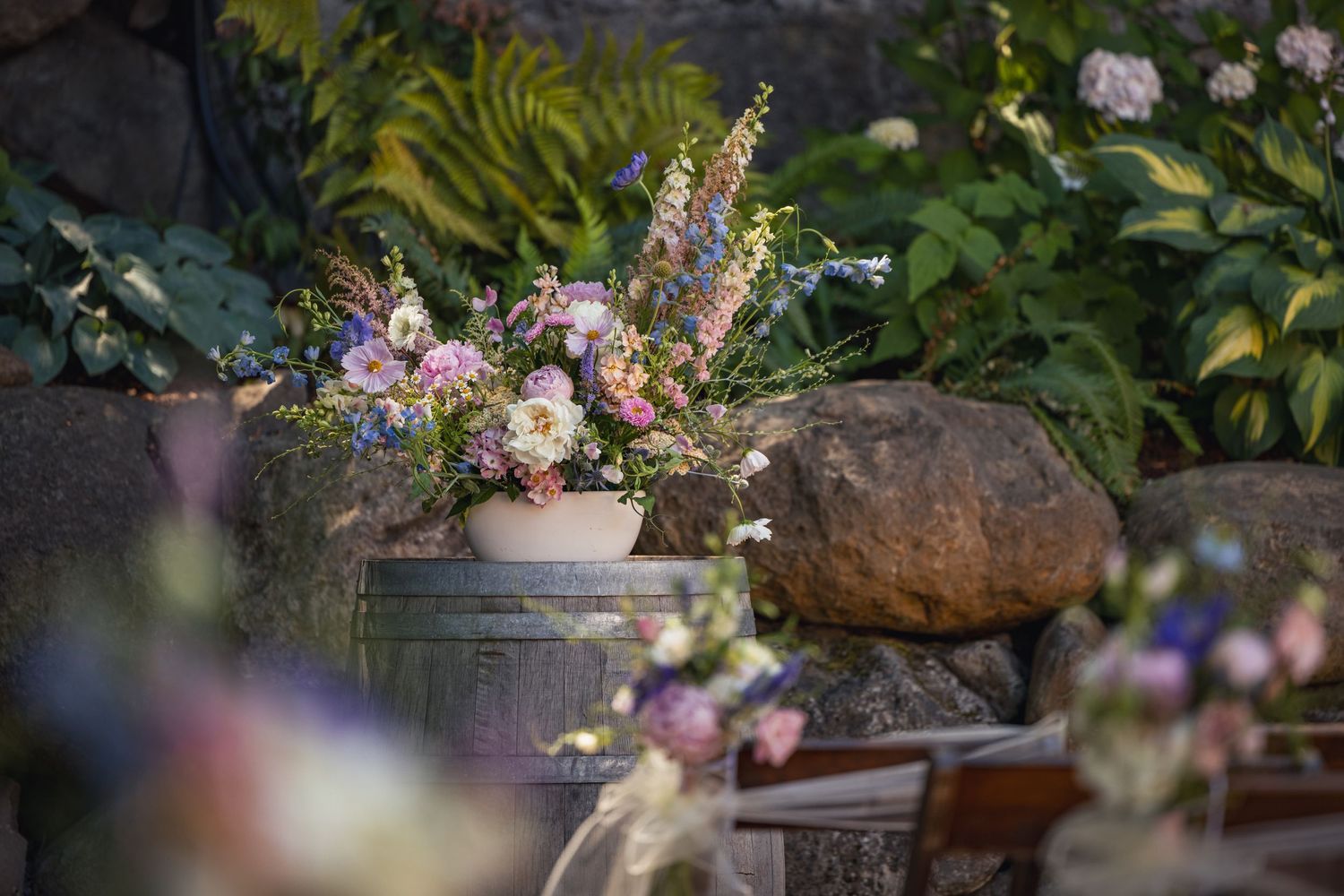 A delicate floral arrangement in a white vase sits among rocks in a garden setting, featuring pink and purple blooms.