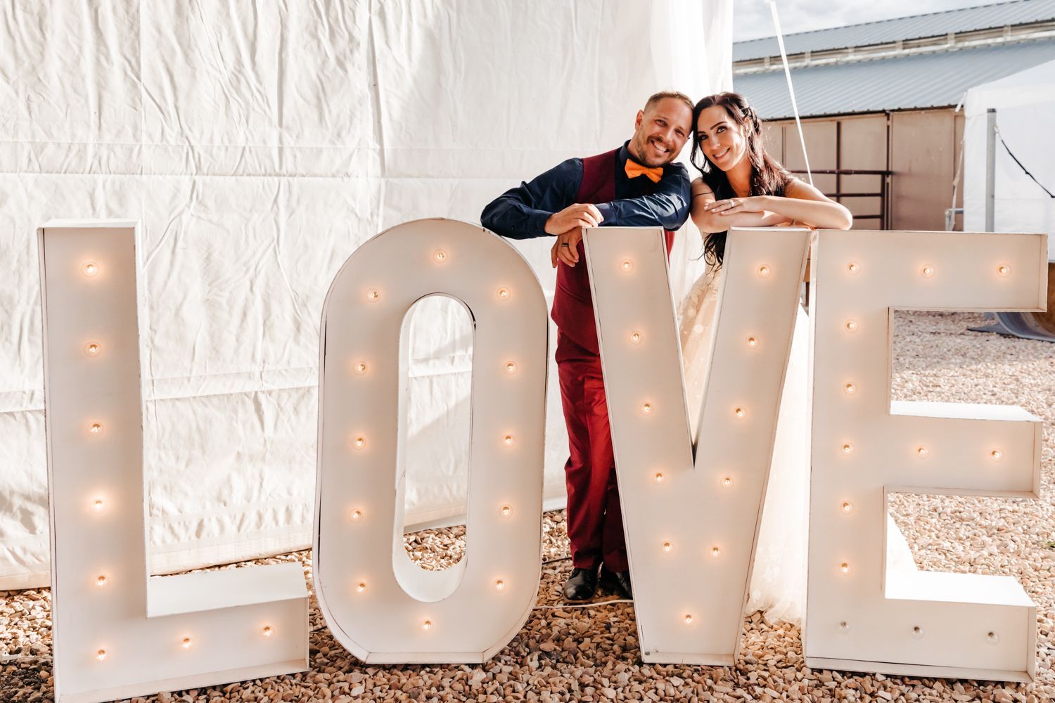 A couple poses affectionately while leaning on large illuminated LOVE letters outdoors in a romantic setting.