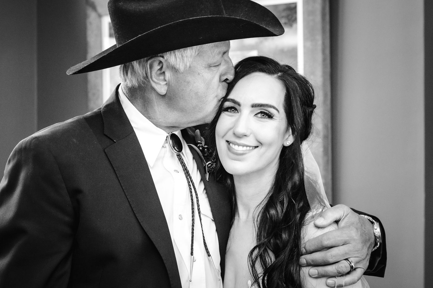 A tender black and white moment showing a sweet kiss on the cheek at what appears to be a wedding celebration.