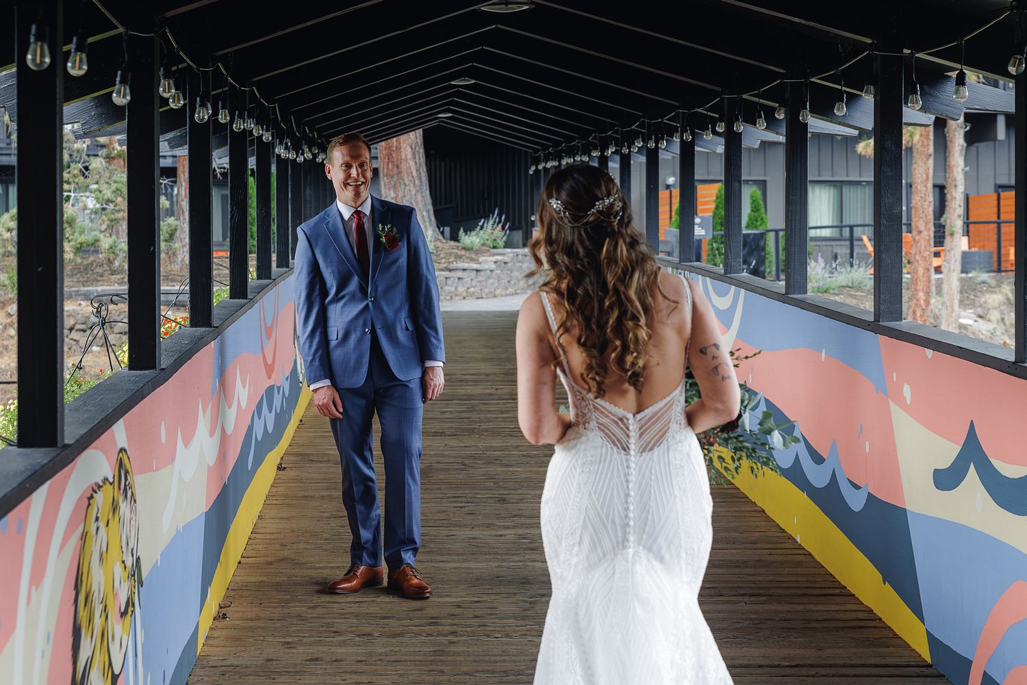 A couple in wedding attire walks under a covered walkway decorated with colorful graffiti art and murals.