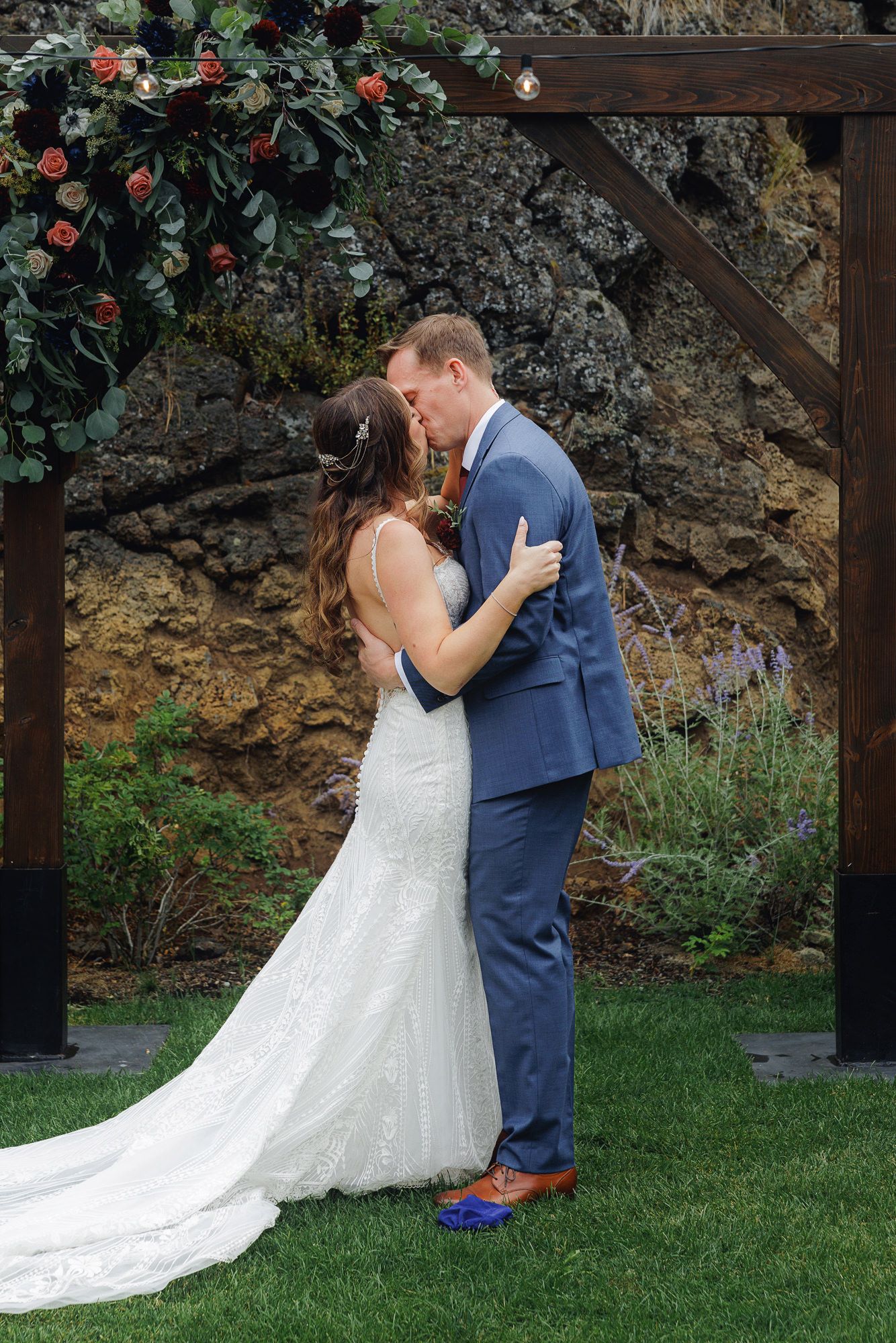 A romantic wedding couple shares an intimate kiss in a garden setting with blooming roses and rustic wooden beams.
