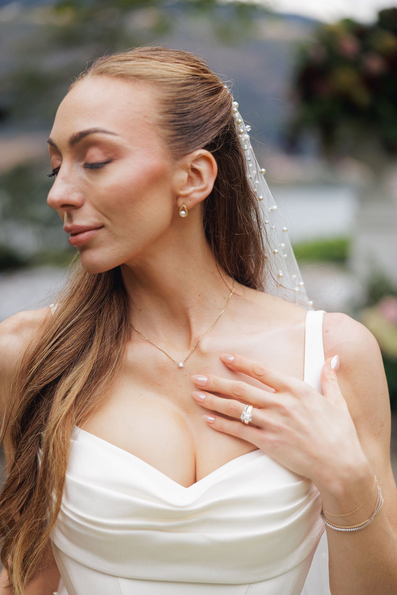 Elegant bride in white gown with pearl veil, diamond ring, and delicate necklace, posing outdoors.