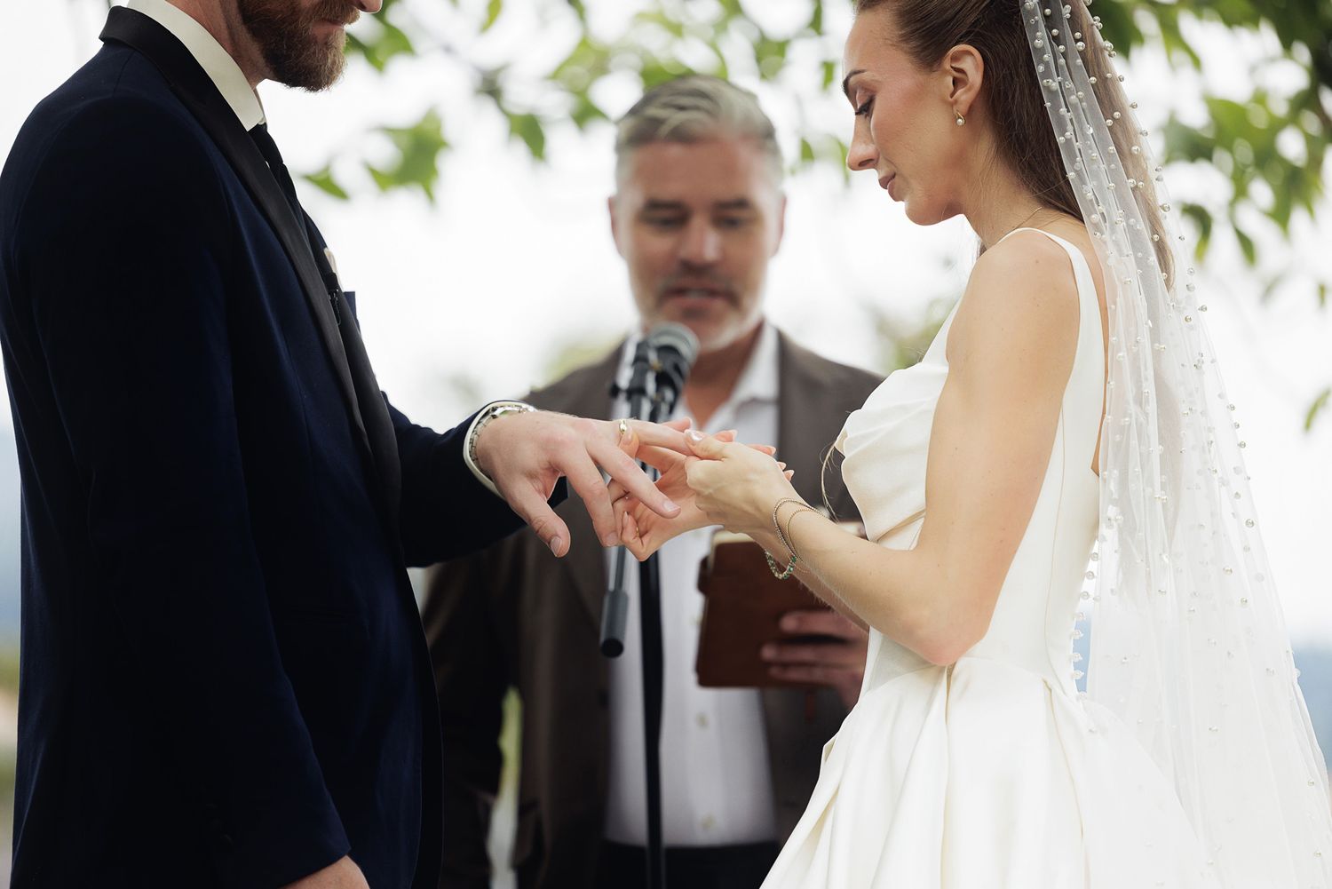 Bride placing wedding ring on grooms finger during outdoor ceremony with officiant in background.