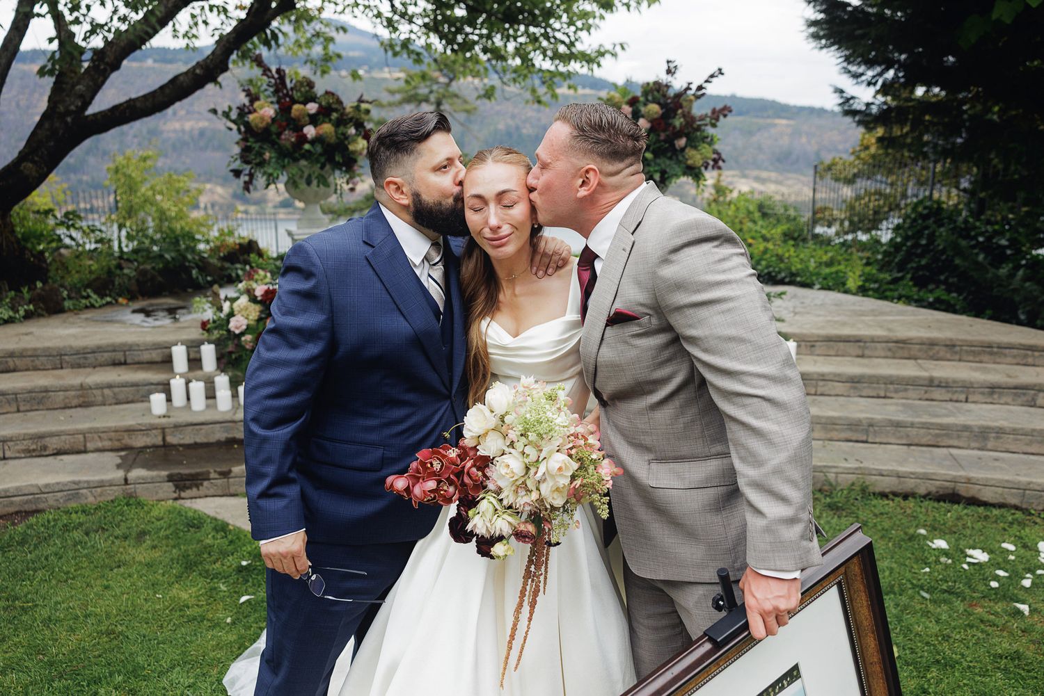Bride in white gown holds bouquet while two men in suits kiss her cheeks at outdoor wedding ceremony.